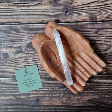 A single-terminated selenite wand with a blunt end and a pointed tip, displayed on a wooden surface, with a business card from 'Arte's Den'.
