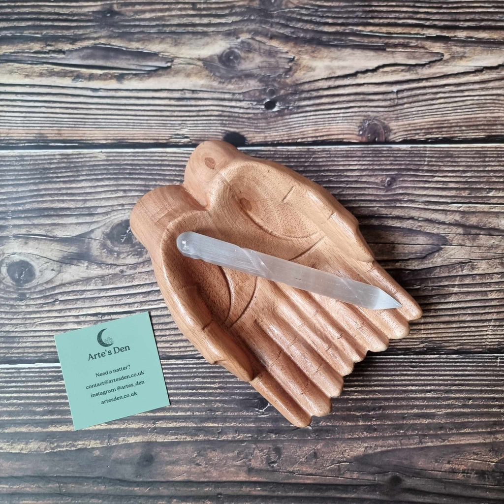 A single-terminated selenite wand with a blunt end and a pointed tip, displayed on a wooden surface, with a business card from 'Arte's Den'.