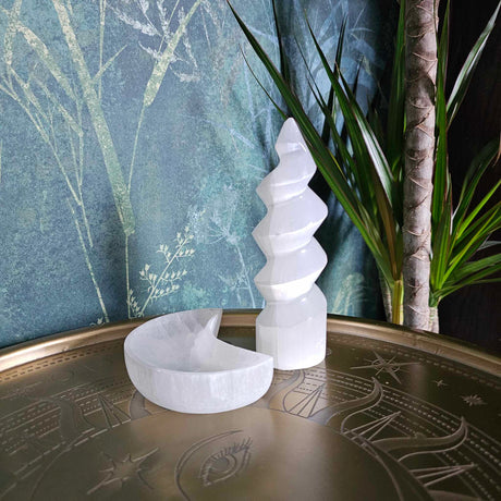 A selenite "unicorn horn" which is a spiral tower next to a selenite crescent moon bowl, on a metal surface with a green plant background