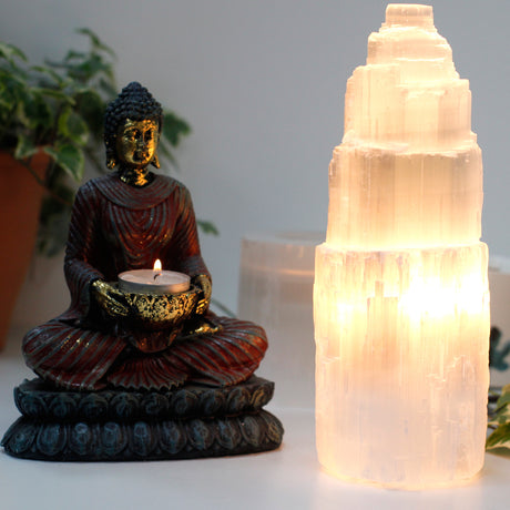 Buddha statue holding a candle next to a glowing selenite crystal tower on a neutral background