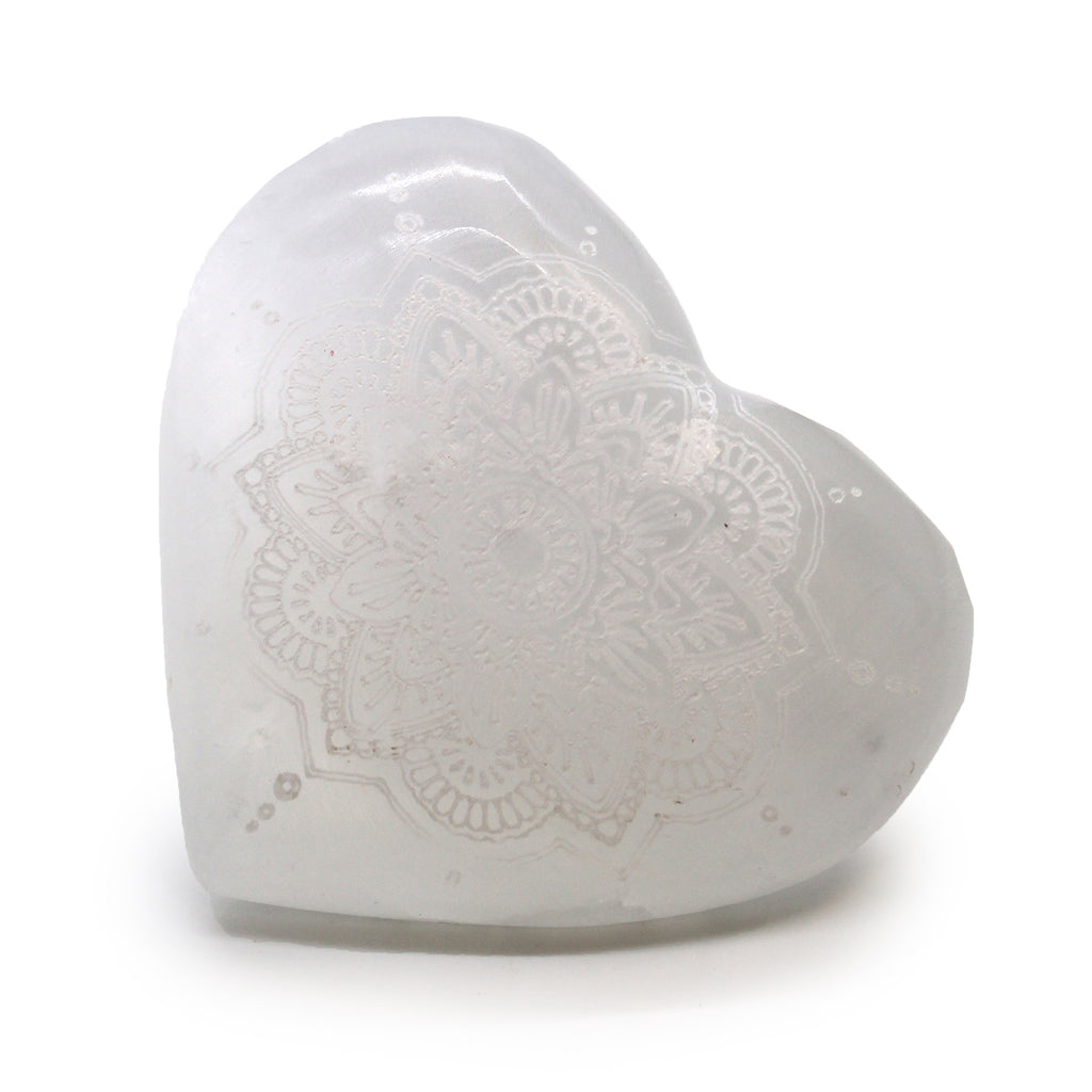 A Selenite heart, carved with intricate mandala design, laid on a white background