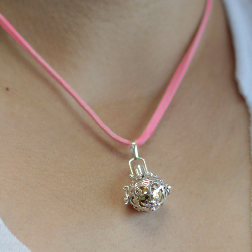 A close-up image of a silver angel bell necklace with a pink necklace, it has a small bell in the centre with a silver and gold design 