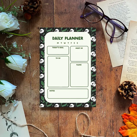 Daily planner on a wooden surface with skulls and plants. Flowers and glasses are on a wooden surface.