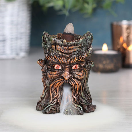 A brown resin Green Man backflow incense burner with smoke cascading downwards, placed on a table with a plant in the background.