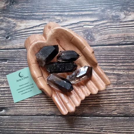 Several Smoky Quartz crystals with a light honey-brown to dark brown color, placed on a wooden palm-shaped dish on a wooden background, with a green Arte's Den business card included.
