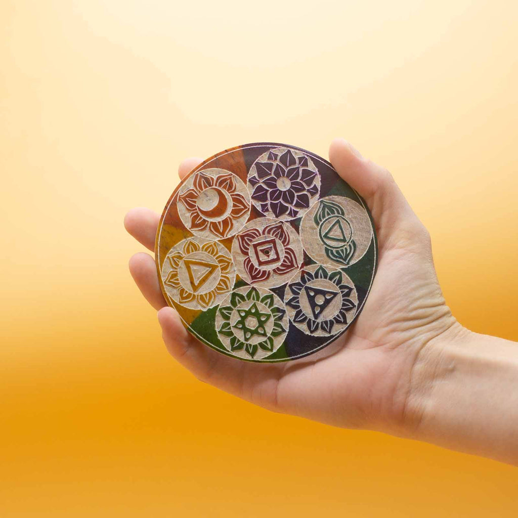 A colourful soapstone incense holder with chakra symbols, held in a hand against a yellow backdrop. 