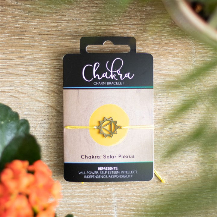 A Solar Plexus Chakra charm bracelet with a yellow band and a silver charm, displayed on a card with 'Chakra' written on it, placed on a wooden surface with orange flowers in the background.