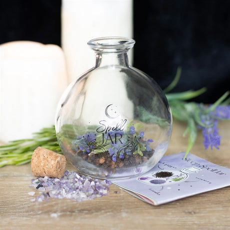 A clear glass spell jar filled with dried flowers and incense, accompanied by a recipe booklet and a candle on a wooden surface.