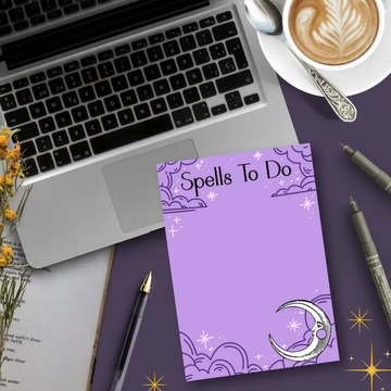 Purple notepad titled 'Spells To Do' on a desk with a laptop, coffee, and stationery.