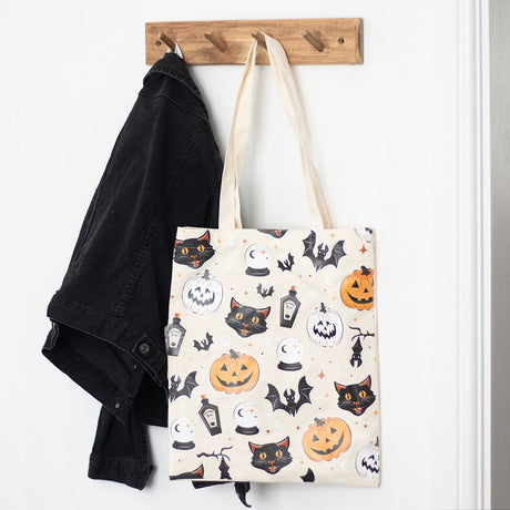 A halloween themed tote bag hanging on a wooden coat hook. It has cats, pumpkins, potions and bats design