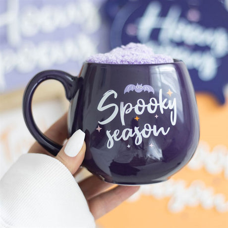 A purple ceramic mug with the text 'Spooky Season' written on it, holding a pair of lilac fluffy socks.