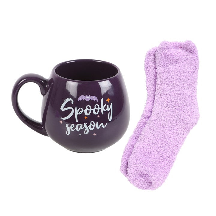 A purple ceramic mug with the text 'Spooky Season' written on it, next to a pair of lilac fluffy socks on a white background