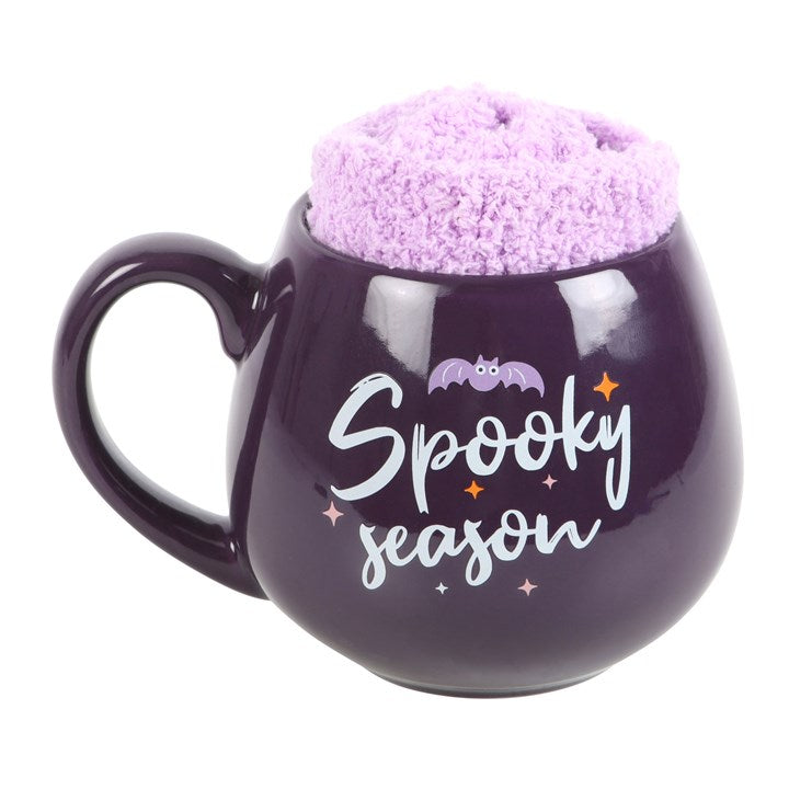 A purple ceramic mug with the text 'Spooky Season' written on it, holding a pair of lilac fluffy socks.