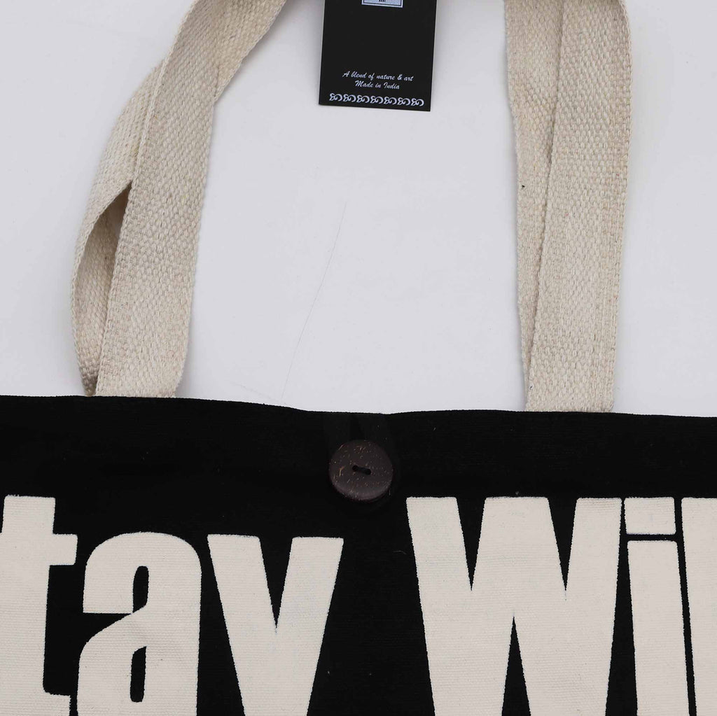 Close-up of a black bag with 'Stay Wild' text, beige handles, and brand tag on a white background