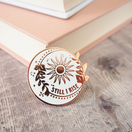 Pin with sun design and 'Still I Rise' text on a wooden surface