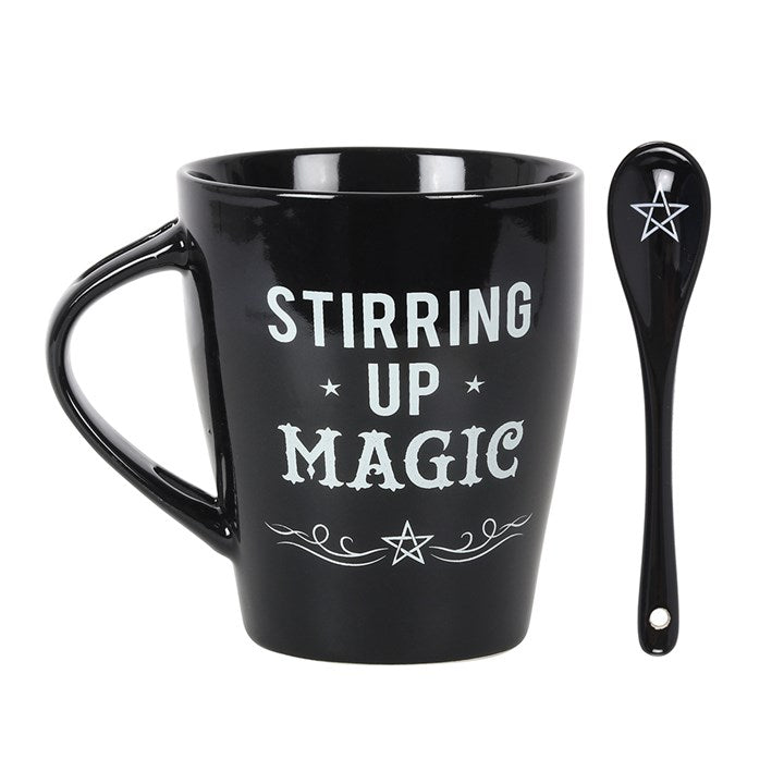 A black mug with a matching black spoon stood beside, with white "Stirring up Magic", swirls and pentagram designs 