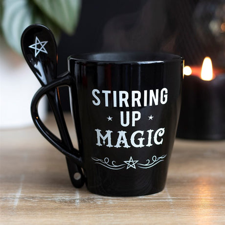 A black mug with a matching black spoon inserted into the handle, with white "Stirring up Magic", swirls and pentagram designs 