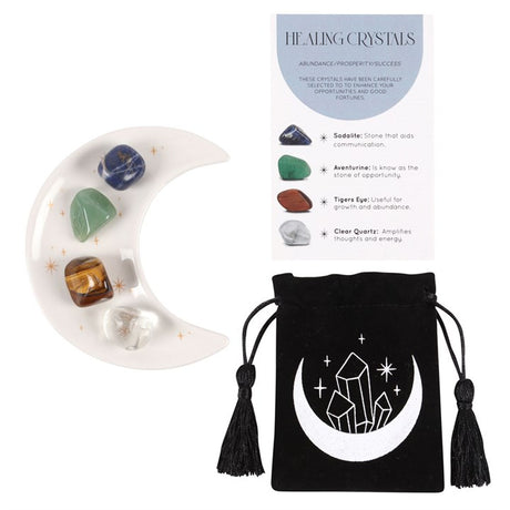 A set of healing crystals displayed with a crescent moon trinket dish, including a green aventurine, a tiger's eye, a clear quartz, and a sodalite stone. The set is packaged in a black drawstring bag with a crescent moon design.