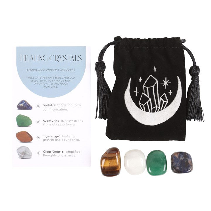 A set of healing crystals displayed with a crescent moon trinket dish, including a green aventurine, a tiger's eye, a clear quartz, and a sodalite stone. The set is packaged in a black drawstring bag with a crescent moon design.