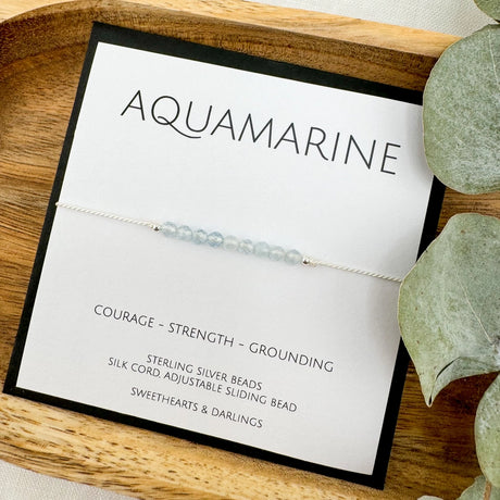 An Aquamarine silk bracelet with beads approximately 3mm in size, displayed on a wooden tray alongside a keepsake card that reads 'Aquamarine - COURAGE - STRENGTH - GROUNDING'