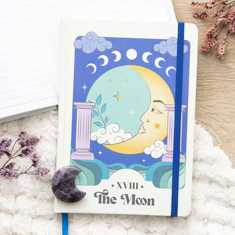 A5 celestial notebook with a design inspired by 'The Moon' tarot card, featuring a moon, moon phases, and clouds motif on the cover alongside an amethyst crescent moon crystal.