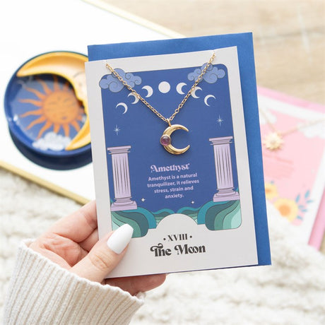 A stainless steel necklace with a crescent moon pendant and an amethyst crystal, displayed on a blue greeting card with tarot card designs and a blue envelope.