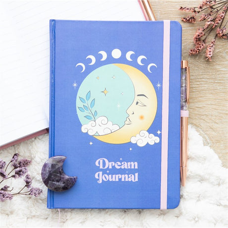 A blue hardcover journal with celestial themed artwork, titled 'Dream Journal', accompanied by an amethyst pen, amethyst crystal moon and decorative dry flowers 