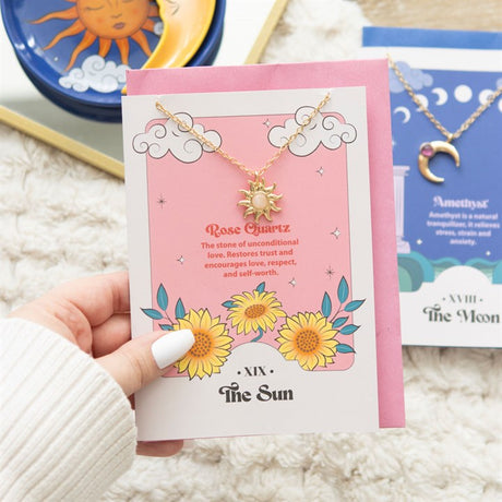 A stainless steel necklace with a sun pendant and a rose quartz crystal, displayed on a pink greeting card with tarot card designs and a pink envelope.