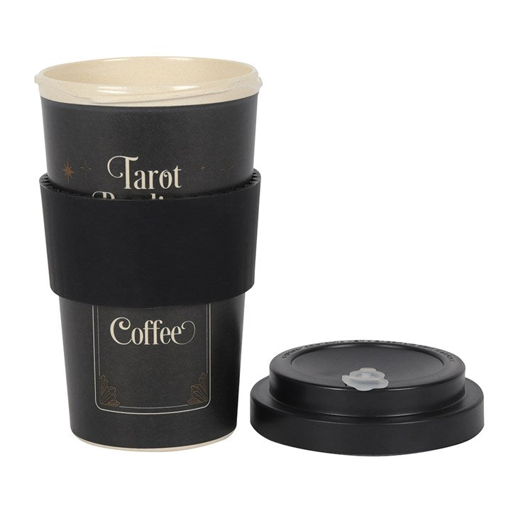 A black bamboo eco travel mug with a 'Tarot Readings' text and a coffee cup design, accompanied by a silicone sleeve and lid which lies next to it on the floor, placed on a table with a candle in the background.
