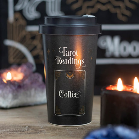 A black bamboo eco travel mug with a 'Tarot Readings' text and a coffee cup design, accompanied by a silicone sleeve and lid, placed on a table with a candle in the background.