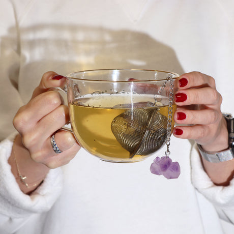 Someone is holding a glass mug with a metal tea strainer in it which has an amethyst crystal on a chain hanging over the side 