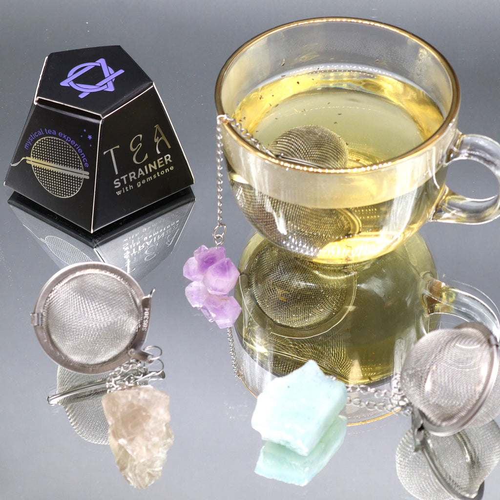 A glass mug with a metal tea strainer in it which has an amethyst crystal on a chain hanging over the side is placed on a mirrored surface with other tea strainers and a box