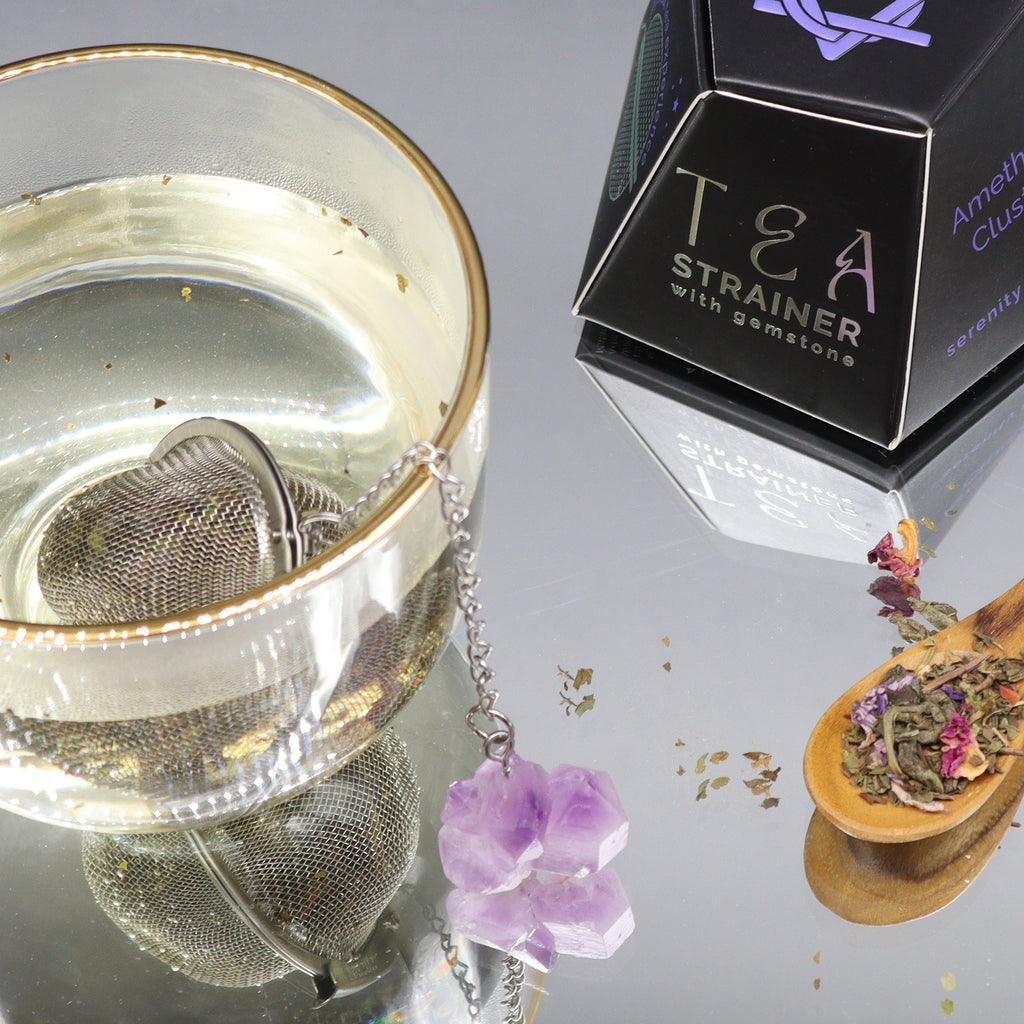 A glass mug with a metal tea strainer in it which has an amethyst crystal on a chain hanging over the side is placed on a mirrored surface with a black box and spoon with herbs.