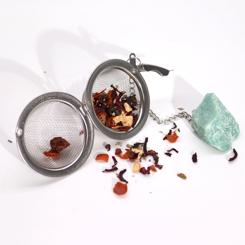 A metal tea strainer with herbs inside, which has a green aventurine crystal on a chain is on a white surface 