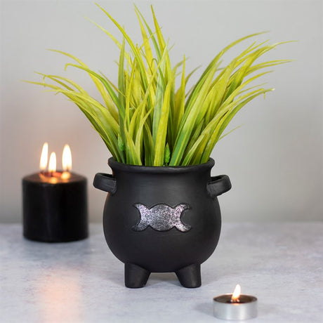 A black terracotta cauldron plant pot with a silver triple moon design, containing green artificial plants, placed on a surface with lit candles in the background.
