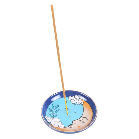 A blue ceramic incense holder with a design resembling the moon and surrounded by clouds. An incense stick is placed in the holder, showing the intended use.