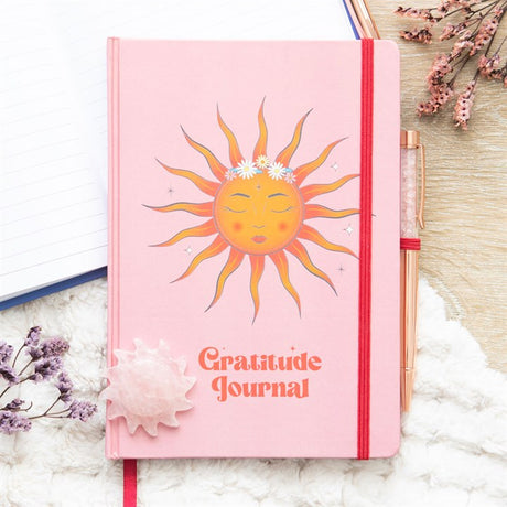 A pink 'Gratitude Journal' with a sun and floral design on the cover, accompanied by a rose quartz pen.
