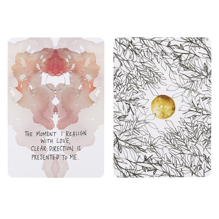 Two oracle cards on a white surface. 
One reads "The moment I realign with love, clear direction is presented to me" and has a pink motif above it. The second card is white but has black line drawings of leaves all over it with a golden orb in the middle.