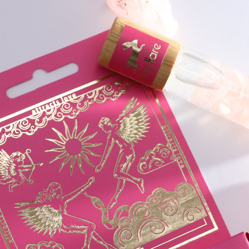 An essential oil gemstone roll-on with a wooden cap by Hop Hare, accompanied by pink tarot card with gold illustrations.