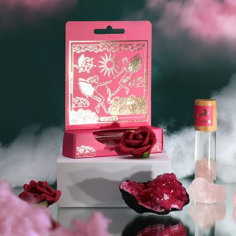 A pink essential oil gemstone roll-on bottle with a decorative cover, placed on a white surface accompanied by a red rose and a small crystal, with a cloud of pink smoke rising in the background.