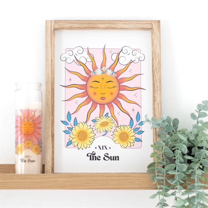 A pink and yellow sun themed artwork and matching tube candle, on a wooden shelf against a white wall.