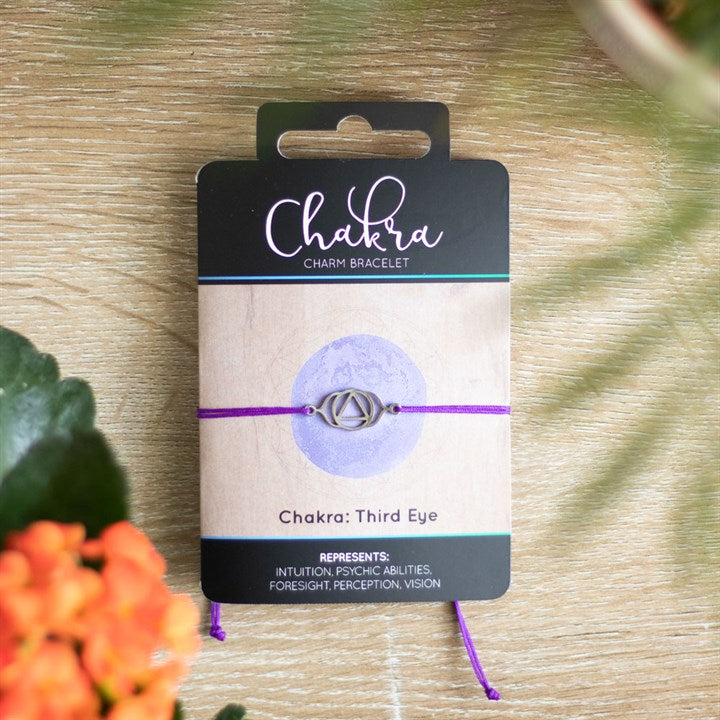 A Third Eye Chakra charm bracelet with a purpley band and a silver charm, displayed on a card with 'Chakra' written on it, placed on a wooden surface with orange flowers in the background.