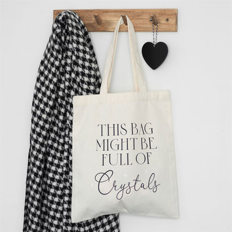 A white cotton tote bag with the text 'This bag might be full of crystals' printed on the front, hanging on a wooden hanger with a black heart-shaped ornament and a black and white jacket