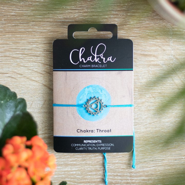 A Throat Chakra charm bracelet with a blue band and a silver charm, displayed on a card with 'Chakra' written on it, placed on a wooden surface with orange flowers in the background.
