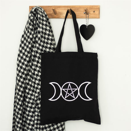 A black cotton tote bag with a triple moon and pentagram design hangs up next to a black and white jacket and black heart 