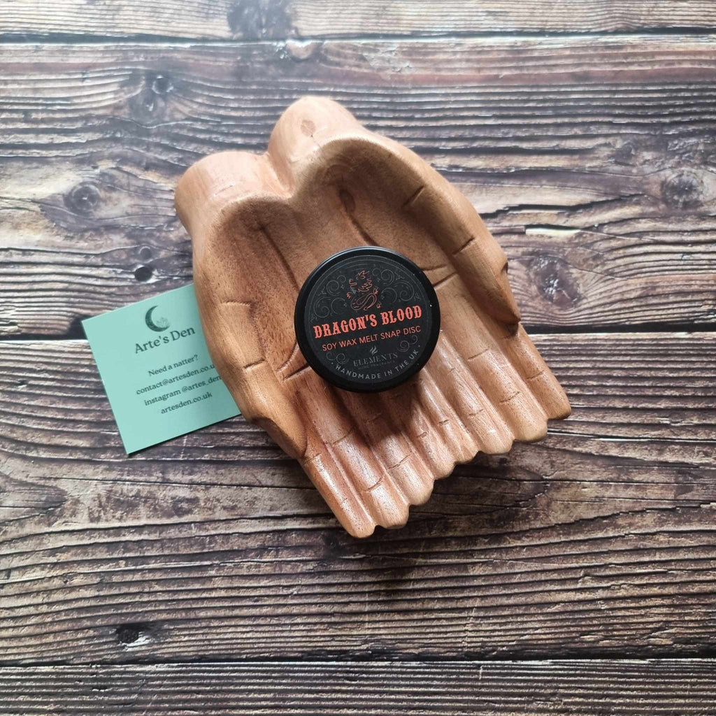 A gothic-themed wax melt tin labelled Dragon's Blood, displayed in a wooden pair of hands on a wooden surface next to a green Arte's Den business card 