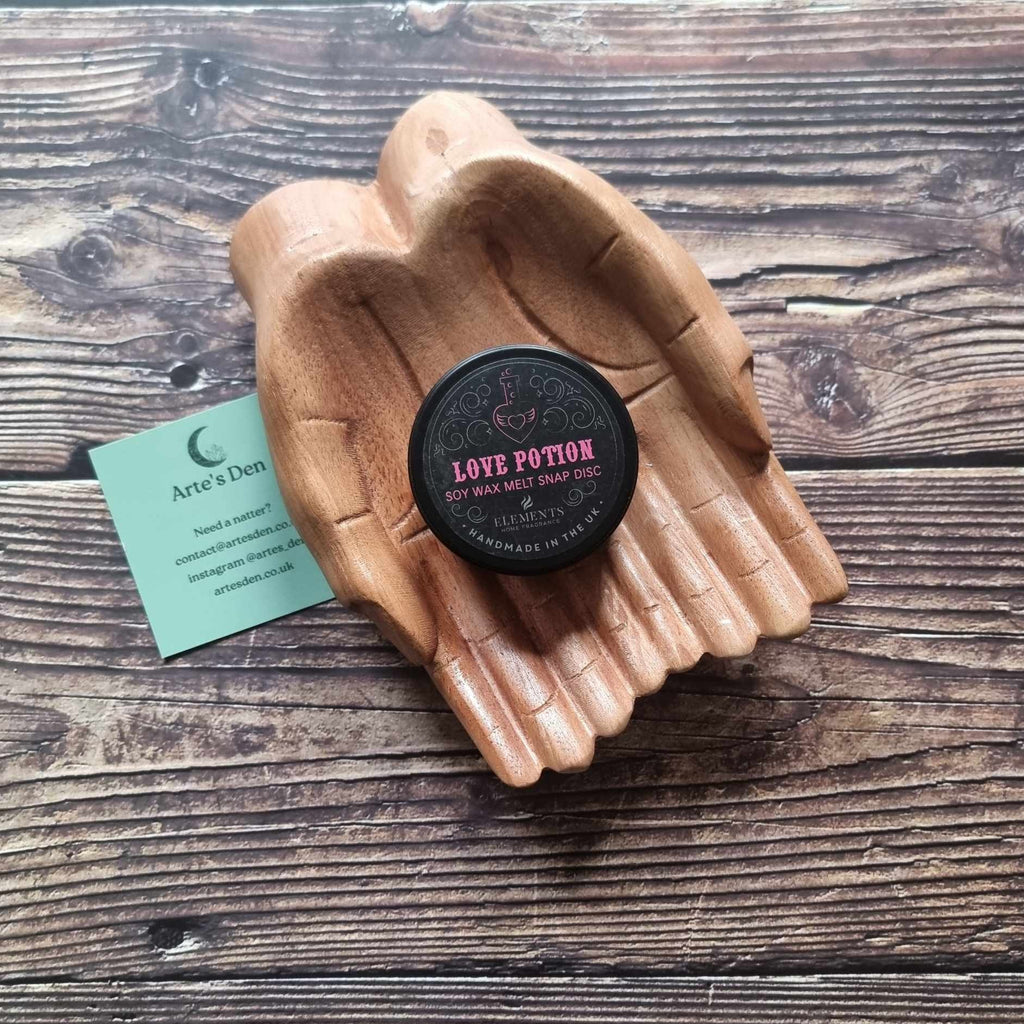 A gothic-themed wax melt tin labelled Love Potion, displayed in a wooden pair of hands on a wooden surface next to a green Arte's Den business card 