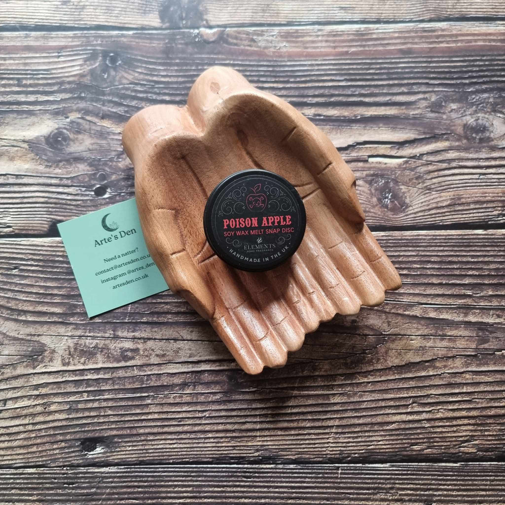 A gothic-themed wax melt tin labelled Poison Apple, displayed in a wooden pair of hands on a wooden surface next to a green Arte's Den business card 