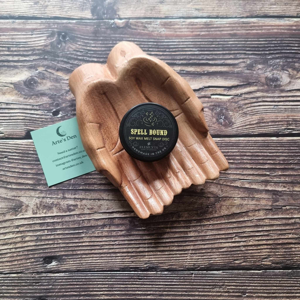 A gothic-themed wax melt tin labelled Spell Bound, displayed in a wooden pair of hands on a wooden surface next to a green Arte's Den business card 