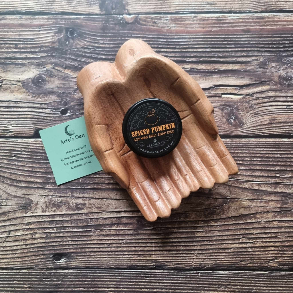 A gothic-themed wax melt tin labelled Spiced Pumpkin, displayed in a wooden pair of hands on a wooden surface next to a green Arte's Den business card 